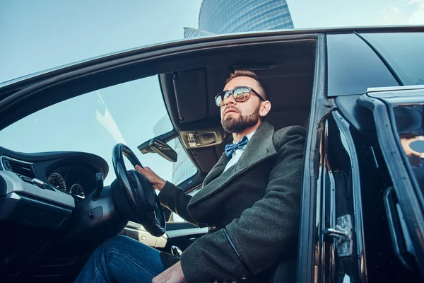 Homem moderno preparado em óculos de sol está sentado em seu carro no estacionamento . — Fotografia de Stock