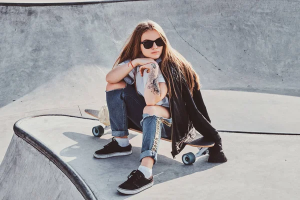 Uzun saçlı ve güneş gözlüklü trendy genç kadın skatepark oturuyor — Stok fotoğraf