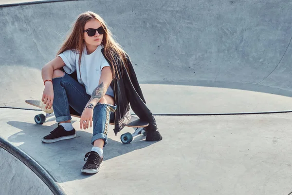 Jovem na moda com cabelos longos e óculos de sol está sentado no parque de skate — Fotografia de Stock