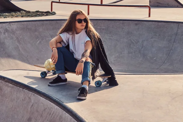 Uzun saçlı ve güneş gözlüklü trendy genç kadın skatepark oturuyor — Stok fotoğraf