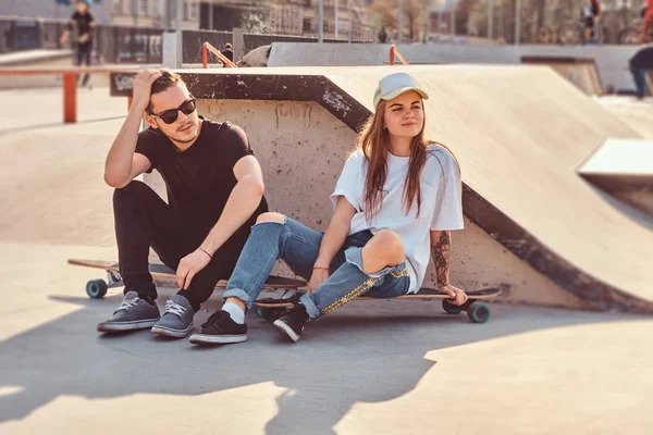 Joven pareja de moda están sentados en el parque de skate soleado con sus longboards — Foto de Stock