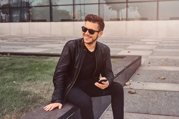 微笑的迷人男人坐在大楼外面边用手机聊天 — 图库照片