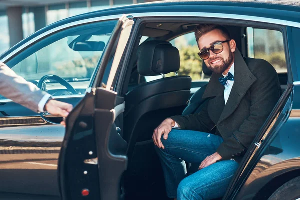 Geschäftiger netter Geschäftsmann mit Sonnenbrille spricht per Smartphone, während sein eleganter Assistent ihm die Tür öffnet. — Stockfoto