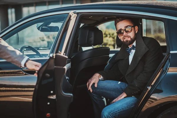 Geschäftiger netter Geschäftsmann mit Sonnenbrille spricht per Smartphone, während sein eleganter Assistent ihm die Tür öffnet. — Stockfoto