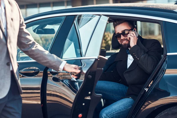 Ocupado agradable hombre de negocios en gafas de sol está hablando por teléfono inteligente, mientras que su elegante asistente está abriendo la puerta para él . — Foto de Stock