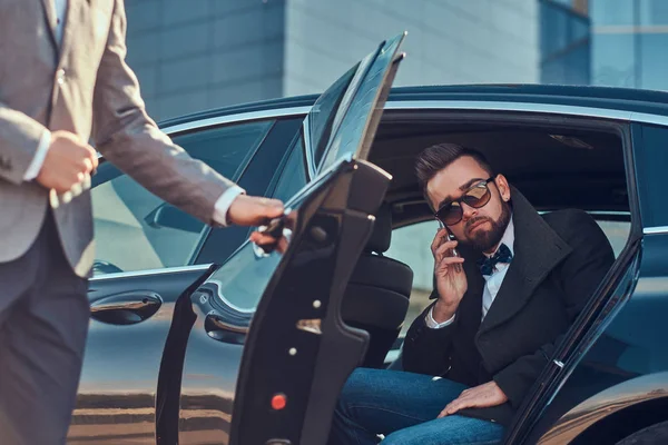 Drukke zakenman in zonnebrillen praat met een smartphone terwijl zijn elegante assistent de deur voor hem opent. — Stockfoto