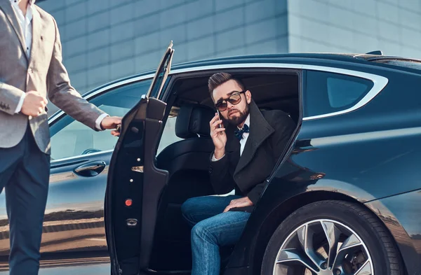 Homem preparado atraente em óculos de sol está falando por smartphone e sentado no carro enquanto seu assistente está abrindo a porta para ele . — Fotografia de Stock