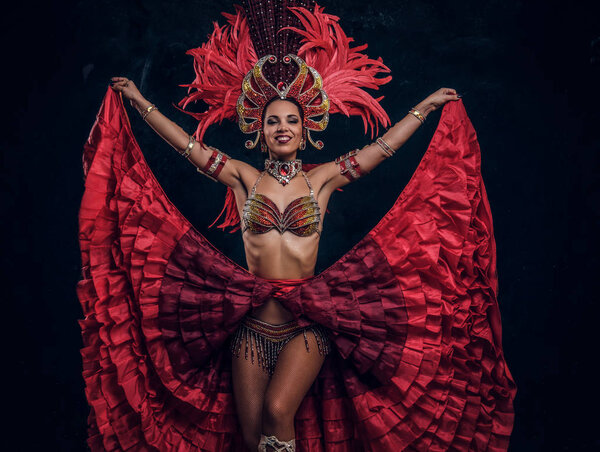 Talented joyful dancer in red feather costume is posing at small dark studio.