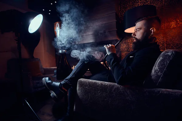 Barbudo hombre relajado está sentado en el sillón y fumar narguile cerca de la chimenea —  Fotos de Stock
