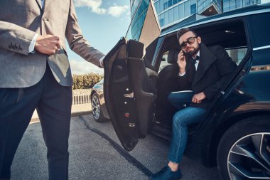 Güneş gözlüğü çekici adam akıllı telefon tarafından konuşuyor ve asistanı onun için kapıyı açarken arabada oturuyor.
