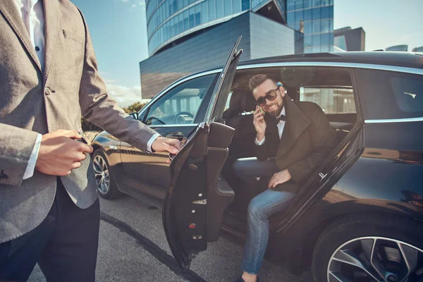 Hombre atractivo en gafas de sol está hablando por teléfono inteligente y sentado en el coche mientras su asistente está abriendo la puerta para él . — Foto de Stock