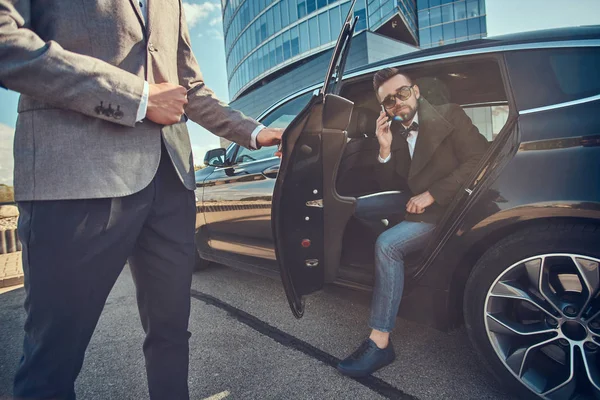 Hombre atractivo en gafas de sol está hablando por teléfono inteligente y sentado en el coche mientras su asistente está abriendo la puerta para él . — Foto de Stock