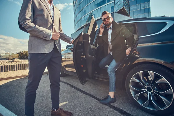 Hombre atractivo en gafas de sol está hablando por teléfono inteligente y sentado en el coche mientras su asistente está abriendo la puerta para él . — Foto de Stock