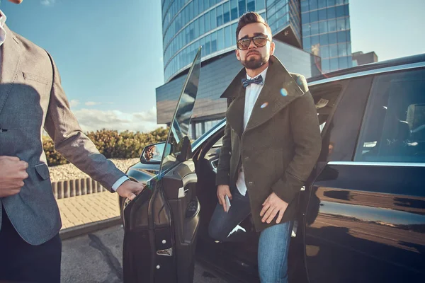 Hombre atractivo en gafas de sol está hablando por teléfono inteligente y sentado en el coche mientras su asistente está abriendo la puerta para él . — Foto de Stock