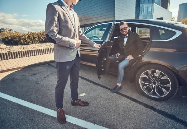 Homme attrayant en lunettes de soleil parle par smartphone et assis dans la voiture tandis que son assistant lui ouvre la porte . — Photo