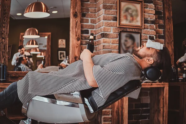 Homem feliz está desfrutando de sua bebida enquanto espera por seu barbeiro na barbearia moderna . — Fotografia de Stock