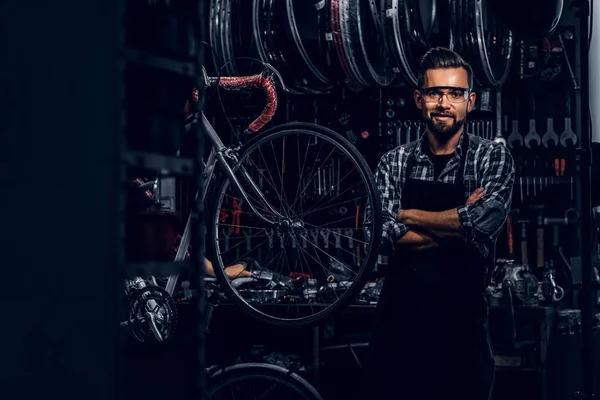 Hombre barbudo guapo en gafas está de pie cerca de la bicicleta fija en su propio taller . — Foto de Stock