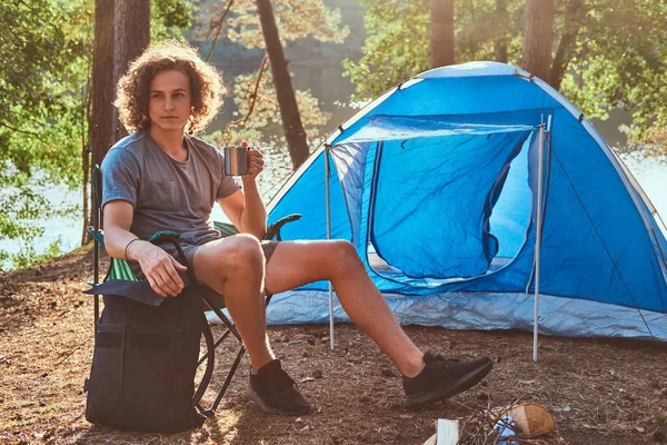 Молодой счастливый путешественник пьет чай возле своей синей палатки. — стоковое фото