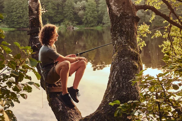 Chico rizado está pescando en el bosque de verano mientras está sentado en el viejo árbol verde —  Fotos de Stock