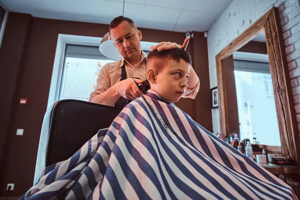 Carino bambino sta ottenendo taglio di capelli alla moda dal barbiere presso il barbiere occupato — Foto Stock