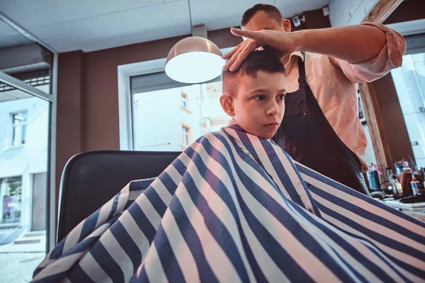 Criança bonito está ficando moderno corte de cabelo de barbeiro na barbearia ocupada — Fotografia de Stock