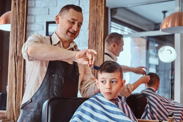 Attractive school boy is getting trendy haircut from mature hairdresser at fashionable hairdressing salon — Stock Photo, Image