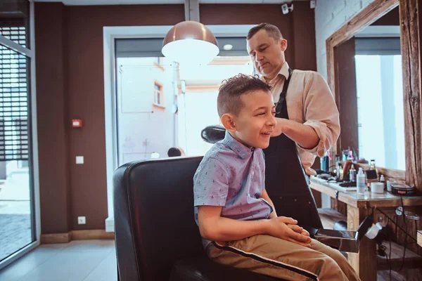 Attractive school boy is getting trendy haircut from mature hairdresser at fashionable hairdressing salon — Stock Photo, Image