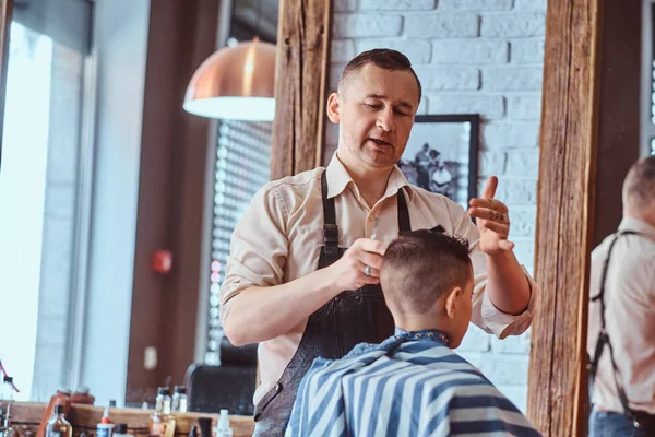 Pequeño chico encantador está consiguiendo corte de pelo de moda de peluquería madura en el salón de peluquería de moda —  Fotos de Stock