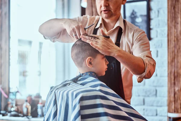 Pequeno menino encantador está ficando moderno corte de cabelo de cabeleireiro maduro no salão de cabeleireiro na moda — Fotografia de Stock