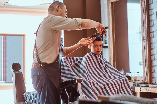 Pequeno menino encantador está ficando moderno corte de cabelo de barbeiro expirienced no salão de cabeleireiro na moda — Fotografia de Stock