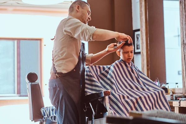 Small charming boy is getting trendy haircut from expirienced barber at fashionable hairdressing salon — Stock Photo, Image