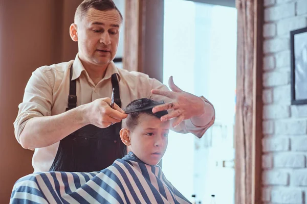 Small charming boy is getting trendy haircut from expirienced barber at fashionable hairdressing salon — Stock Photo, Image