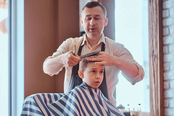 Small charming boy is getting trendy haircut from expirienced barber at fashionable hairdressing salon — Stock Photo, Image