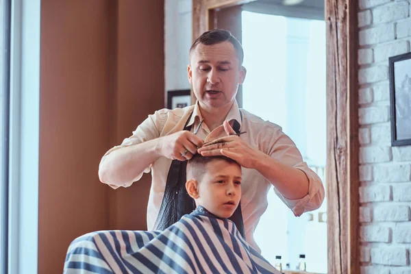 Small charming boy is getting trendy haircut from expirienced barber at fashionable hairdressing salon — Stock Photo, Image
