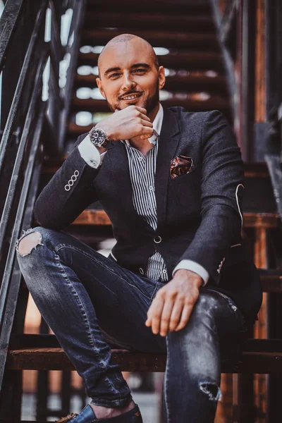 Retrato del hombre sonriente en chaqueta y pantalones vaqueros rasgados que está sentado en las escaleras . — Foto de Stock