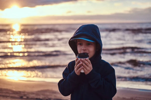 Menino está bebendo chá à beira-mar, na hora do pôr do sol — Fotografia de Stock