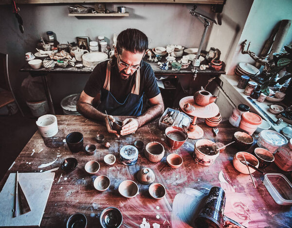 Entusiastici potter in glasses at his own workshop is workig on a new handmade teapot.