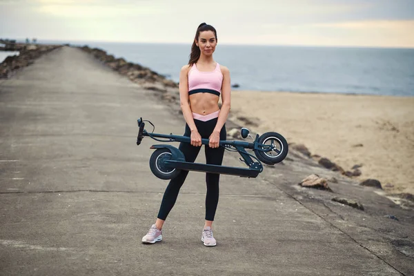 Woman in activewear is holding folded electro scooter