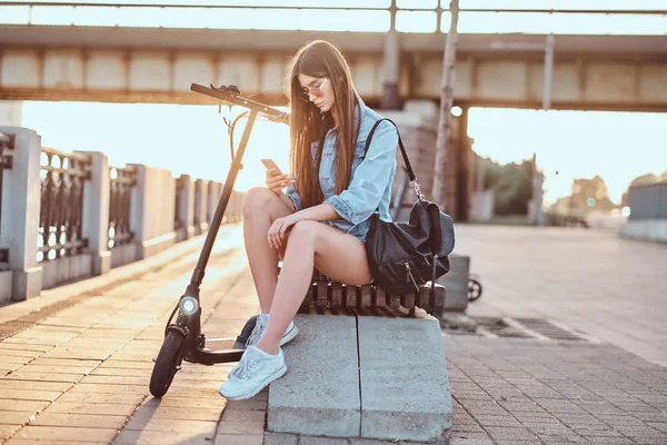 Красивая девушка сидит на скамейке со своим скутером — стоковое фото