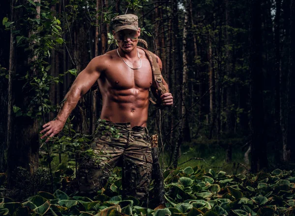 Young handsome soldier with naked torso is walking in the green deep forest at hot summer day. — Stock Photo, Image