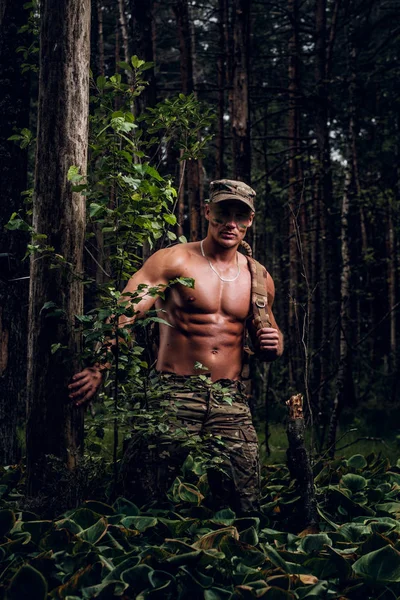 Junger schöner Soldat mit nacktem Oberkörper spaziert an heißen Sommertagen im grünen tiefen Wald. — Stockfoto