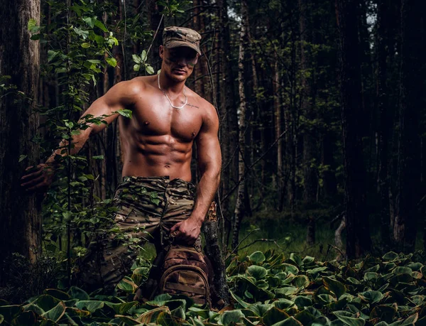 Jovem soldado bonito com tronco nu está andando na floresta profunda verde no dia quente de verão . — Fotografia de Stock