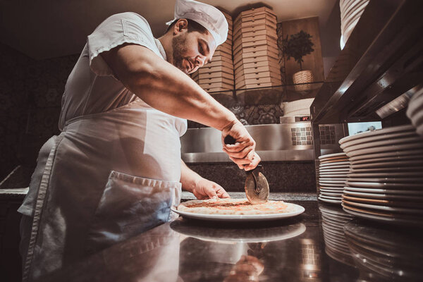 Chef is cutting tasty pizza with special knife at the kitchen.