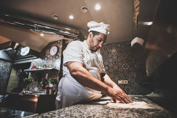 Italian chef in uniform is preparing pastry for pizza at the kitchen.