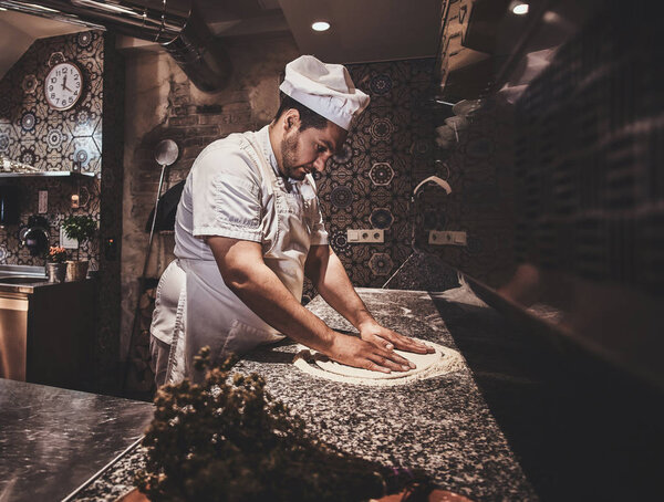 Italian chef in uniform is preparing pastry for pizza at the kitchen.