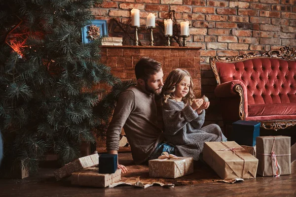 Cheerful girl is holding christmas decoration ball while sitting with her father on the floor, surrounded with gifts. — Stock Photo, Image