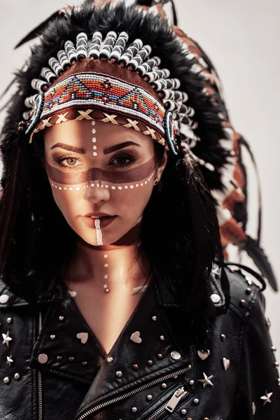 Confident young woman posing in a studio wearing tribal headdress and make-up, wearing leather coat — Stock Photo, Image