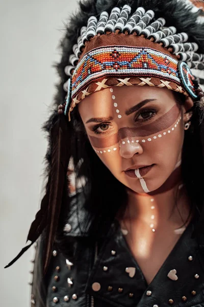 Confident young woman posing in a studio wearing tribal headdress and make-up, wearing leather coat — Stock Photo, Image