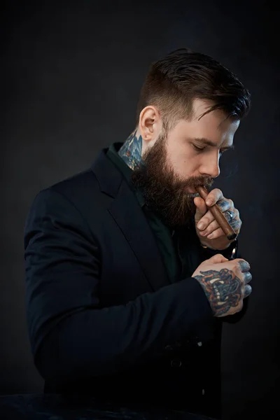 Elegantemente vestido hombre barbudo con tatuajes en el cuello y la mano enciende un cigarro —  Fotos de Stock