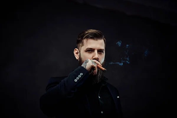 Brutal young businessman smokes a cigar in a dark studio. — Stock Photo, Image